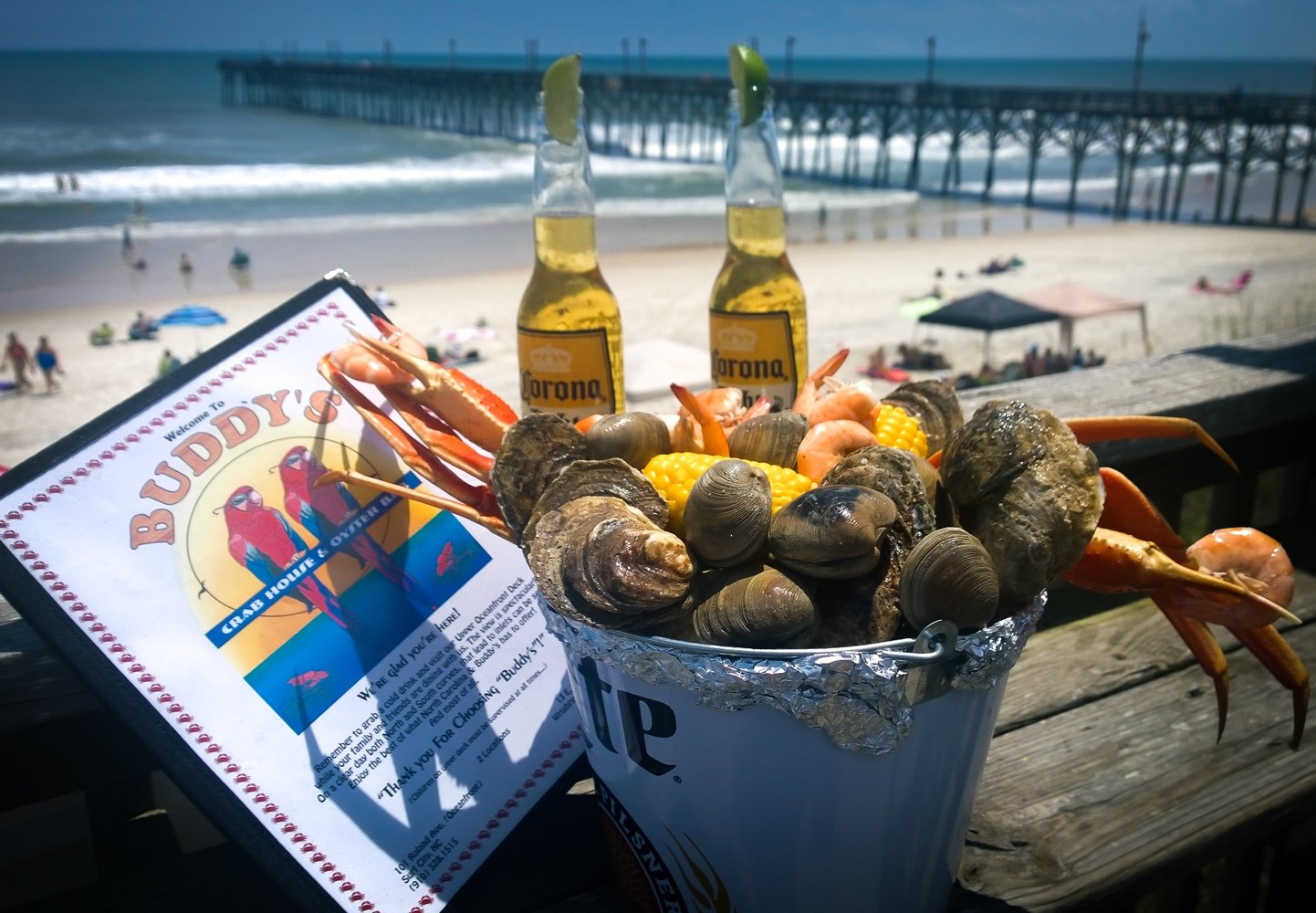 A steamer bucket of crab legs, clams, shrimp and corn on the deck rail, with the Surf City Pier behind it